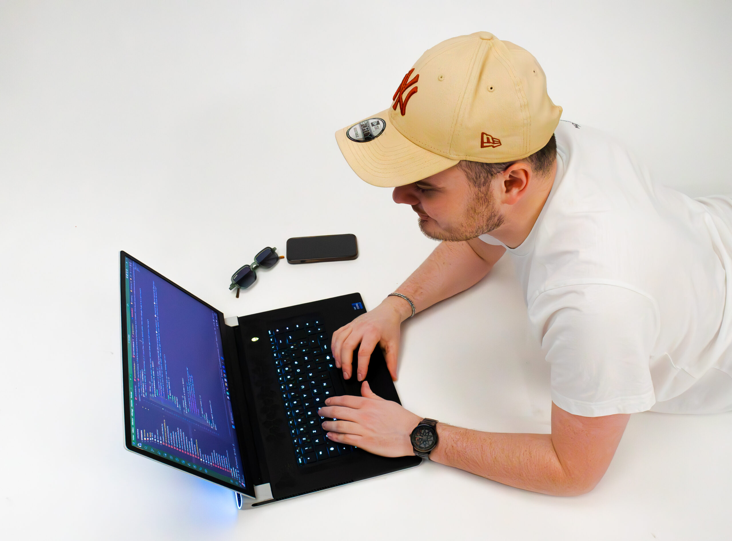 Photo lifestyle d’un développeur web concentré sur son laptop, vue plongeante, avec smartphone et lunettes sur fond blanc.